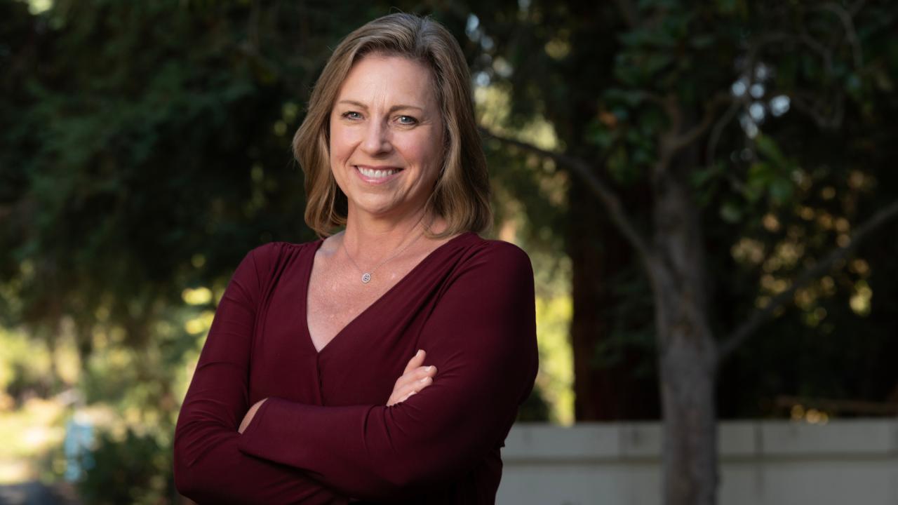 in an outdoor setting on a sunny day, a woman wearing a maroon sweater, has her arms crossed and is smiling at the camera.
