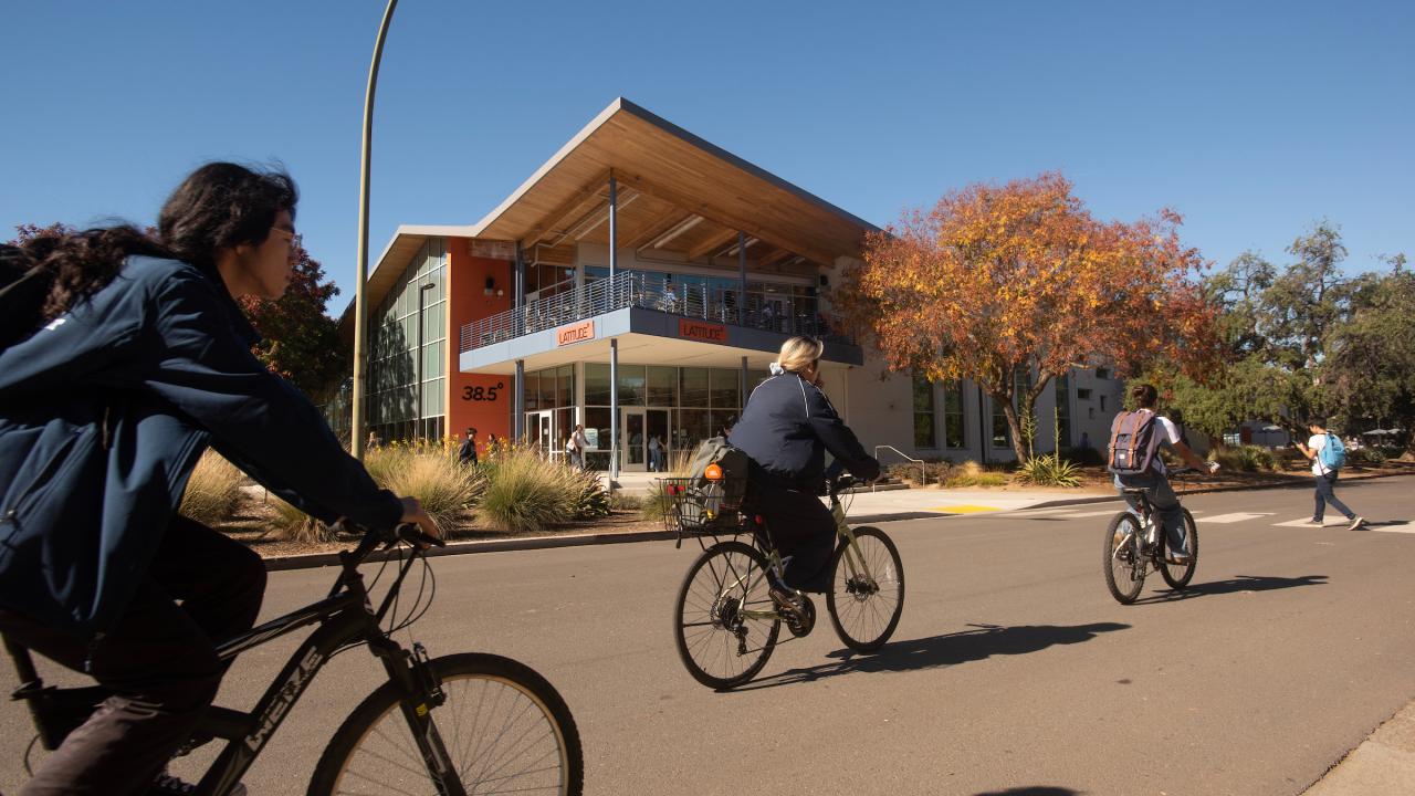 On a sunny fall day, three students on bicycles ride along a path with the restaurant Latitude in the background.