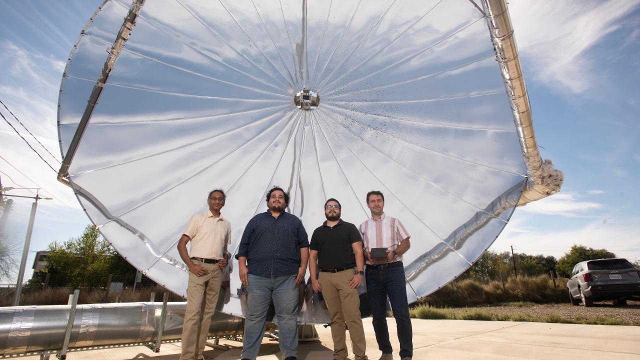 Four men stand outdoors in front of a large circular mirrored dish. 