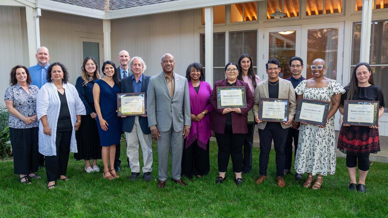 Group photo on lawn of diverse adults holding framed certificates outside a lighted building