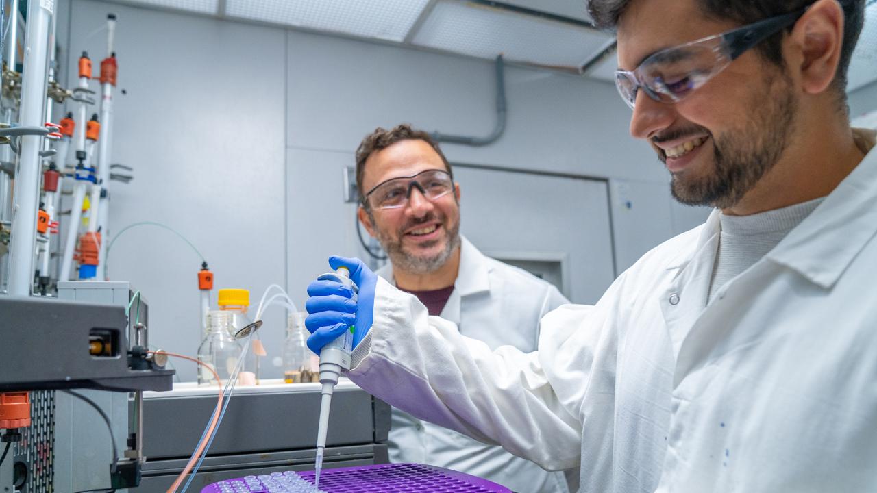Two scientists in lab coats and goggles smiling while pipetting at a lab bench