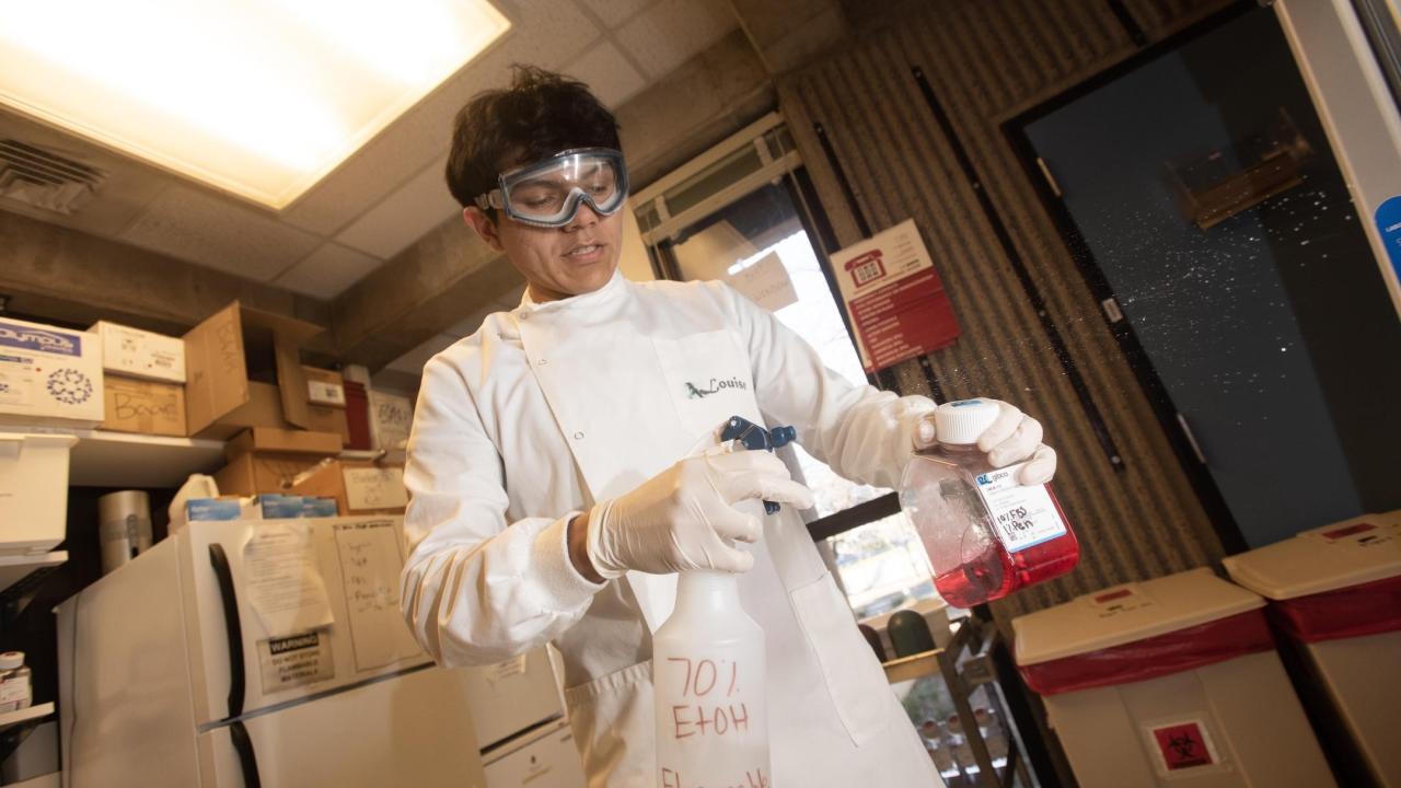 Undergraduate researcher wears safety goggles and gloves pours red liquid into a spray bottle labeled “70% EtOH Flammable” inside a laboratory workspace with shelves and equipment in the background.