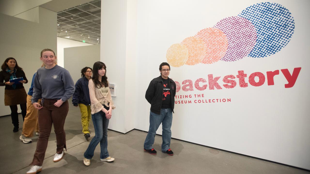 Visitors walking past a colorful exhibition wall labeled "Backstory" in a gallery.