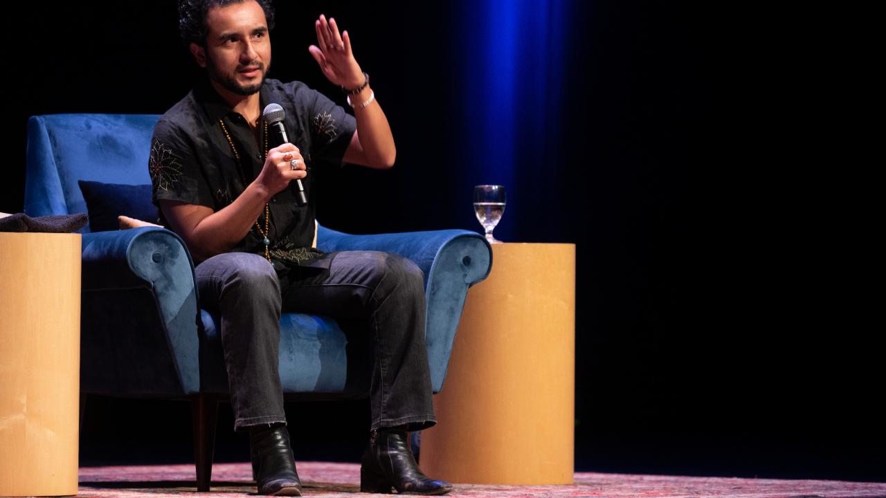 A speaker, Javier Zamora, gestures while seated on a blue chair, water glass nearby, under stage lights.