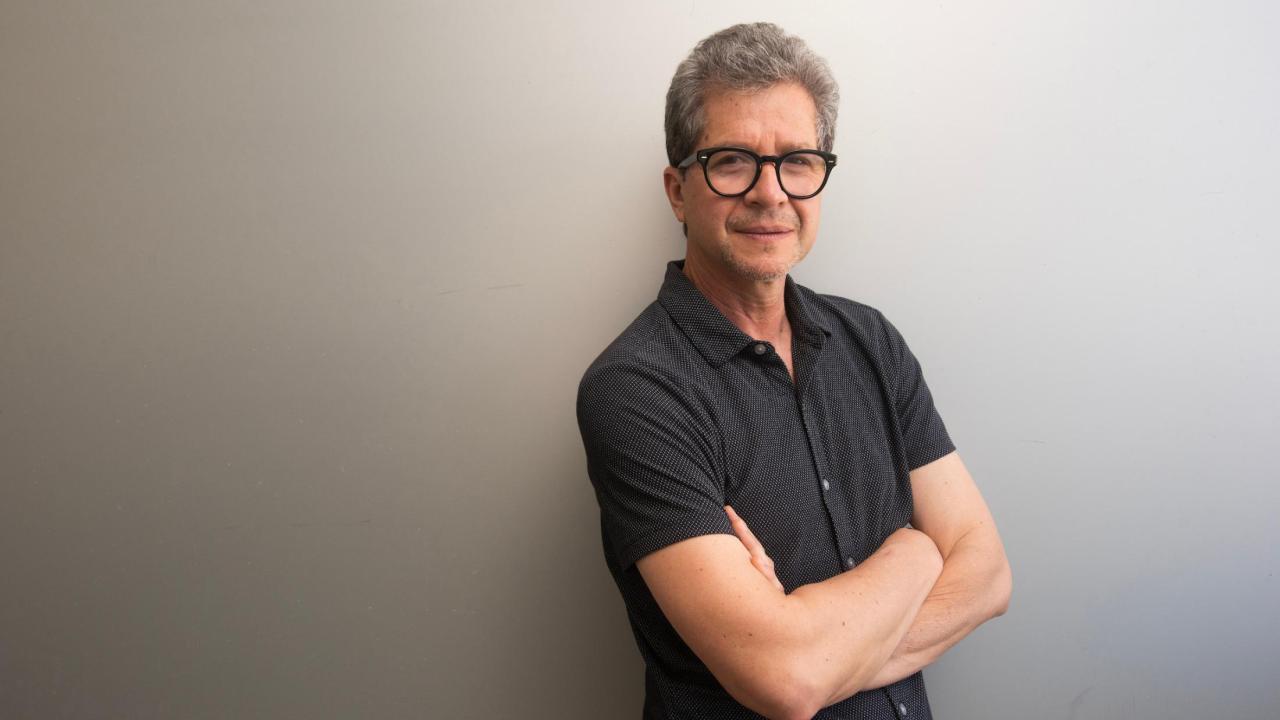 Smiling middle-aged man with glasses, arms crossed against a light gray wall.