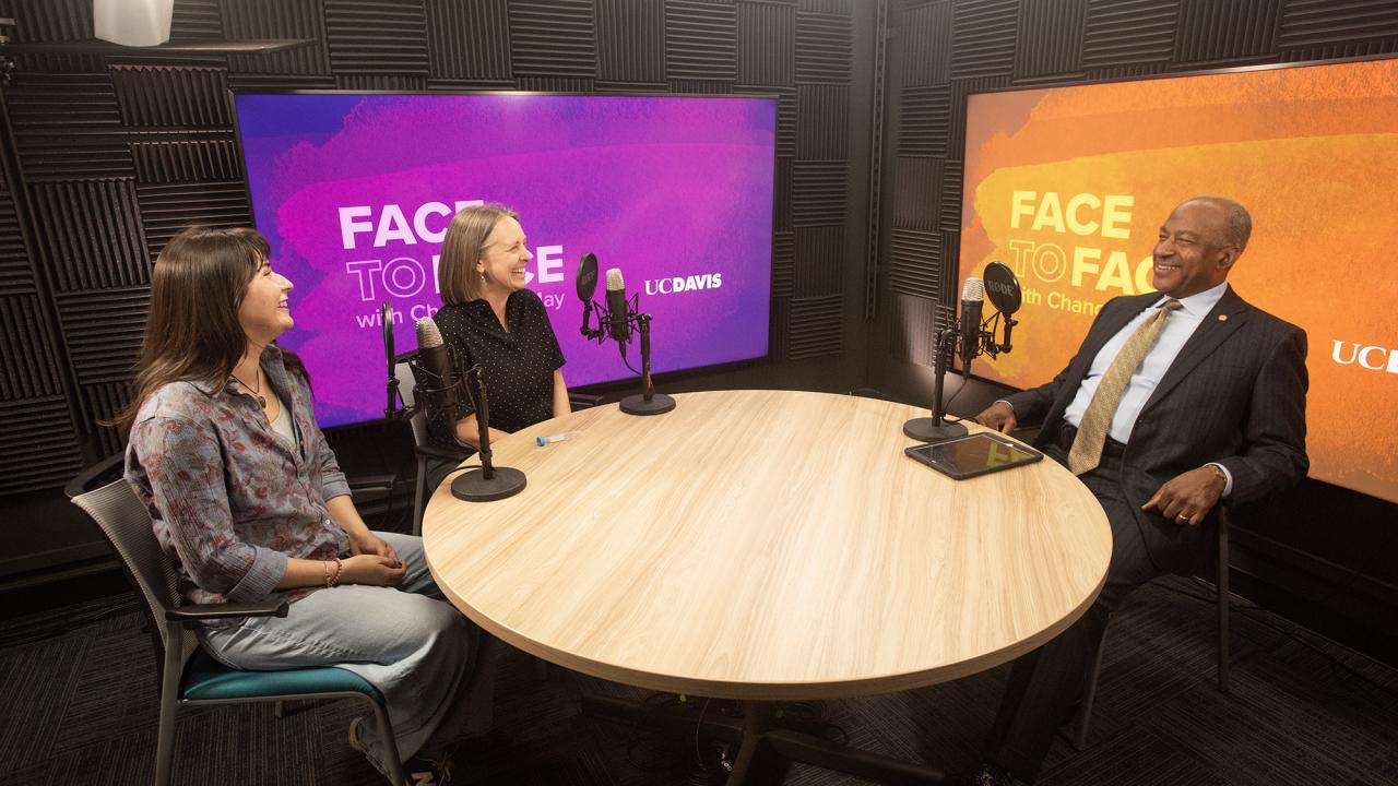 Studio podcast with three people at round table, mics, purple and orange backdrops