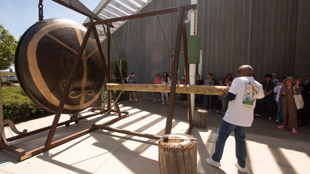 Outdoor scene: man swinging mallet at large bronze sculpture while crowd watches under awning