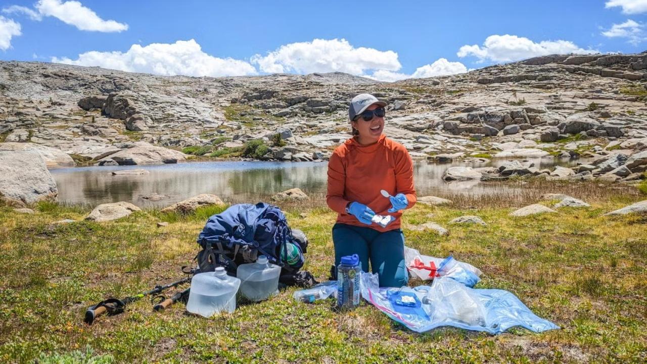 Person in orange jacket kneeling with camping gear beside a small alpine lake under blue sky