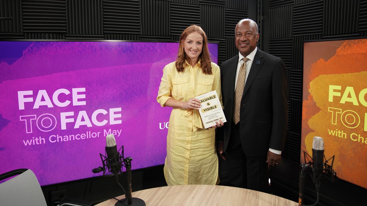 Two people on 'Face to Face' set; woman in yellow holds certificate, man in suit.