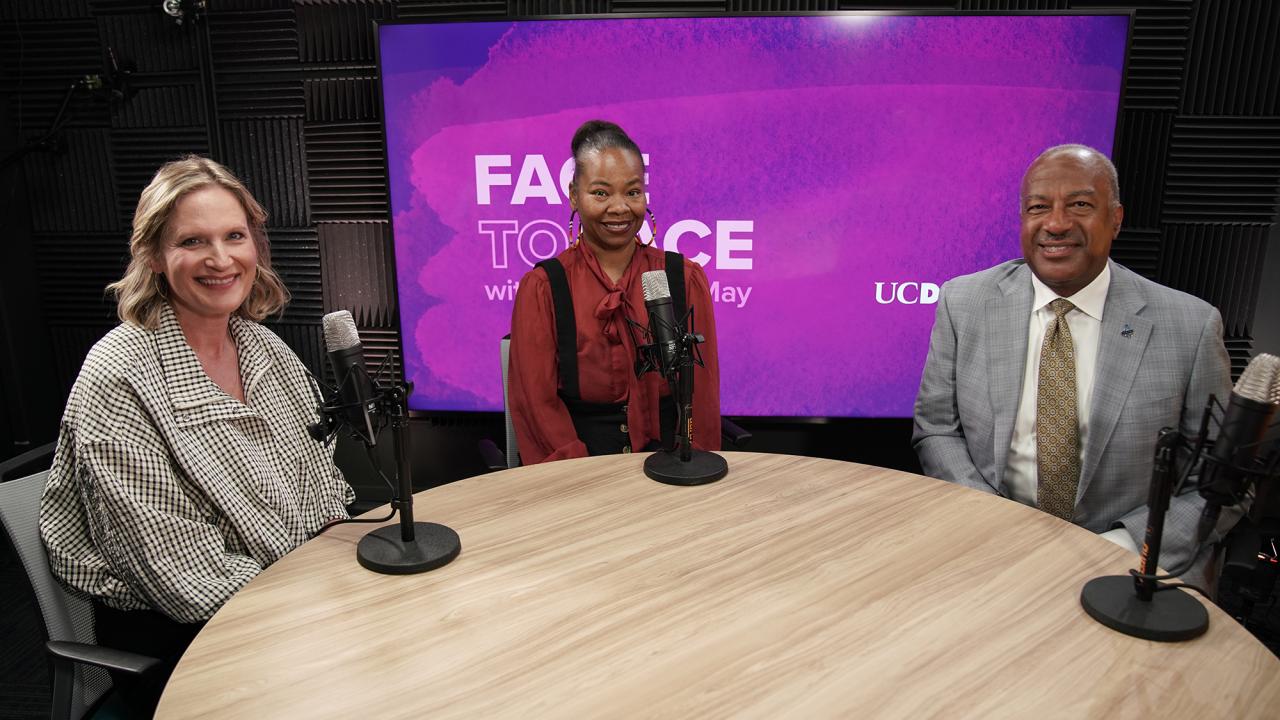 Three smiling people seated at a round podcast table with microphones and purple backdrop