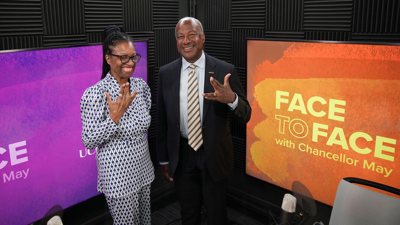 Two smiling presenters in studio beside orange 'Face to Face' set screen