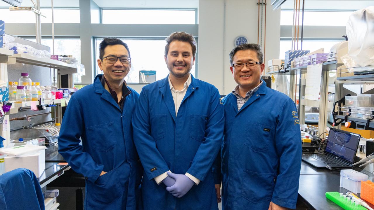 Three scientists in blue lab coats stand smiling in a laboratory filled with equipment.
