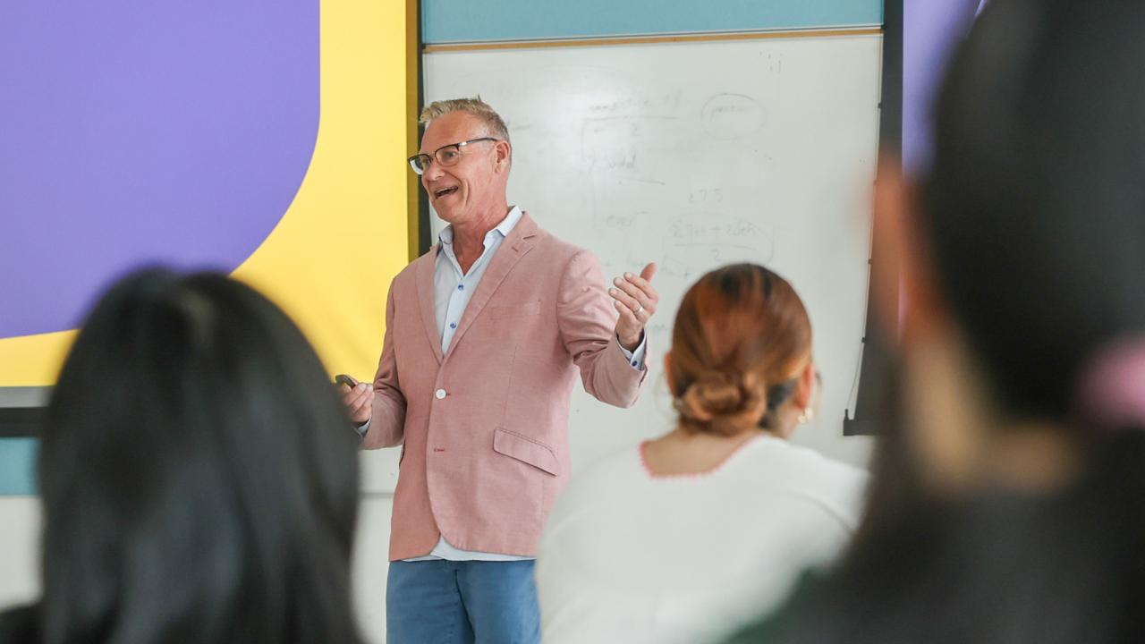Man in pink blazer lecturing at a whiteboard to seated students