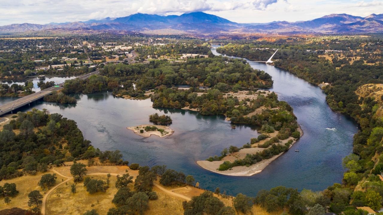 Aerial photo of meandering Sacramento River with tree-covered islands and distant mountains and white sundial bridge