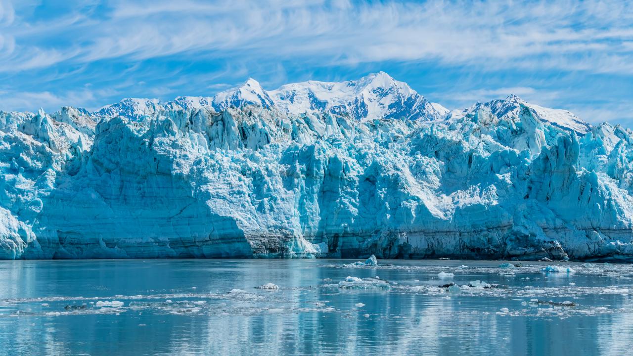 Blue glacier wall with snow-capped peaks reflecting on calm icy water