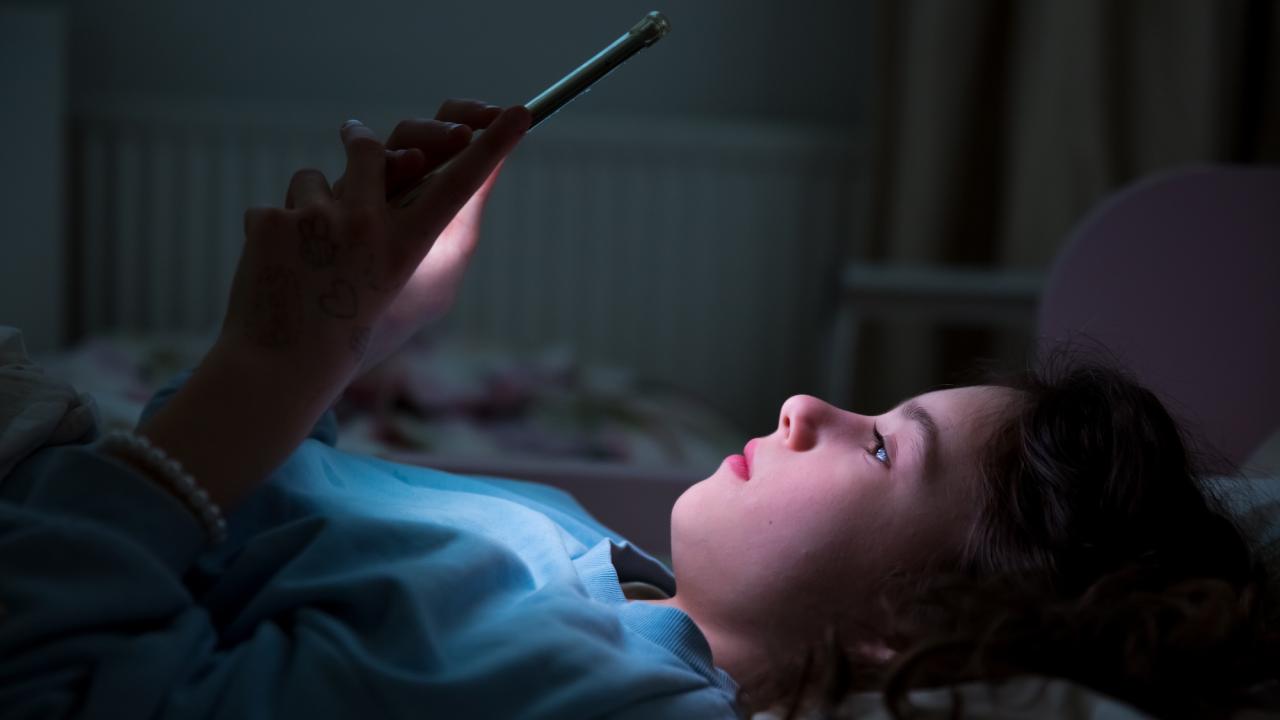 Girl lying in dark bedroom, face glowing from smartphone screen. UC Davis researchers have created a new public database that tracks bills regulating digital media for minors. (Adobe Stock)