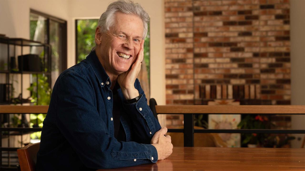 Smiling man in blue shirt leaning on hand at wooden table in kitchen