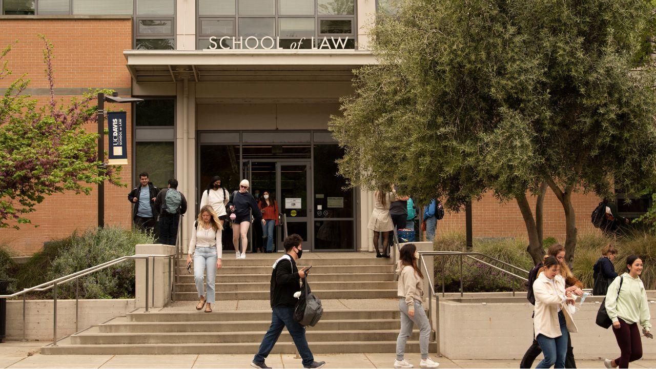 Students walk out of a tree-lined brick building with the words "School of Law" over the door.