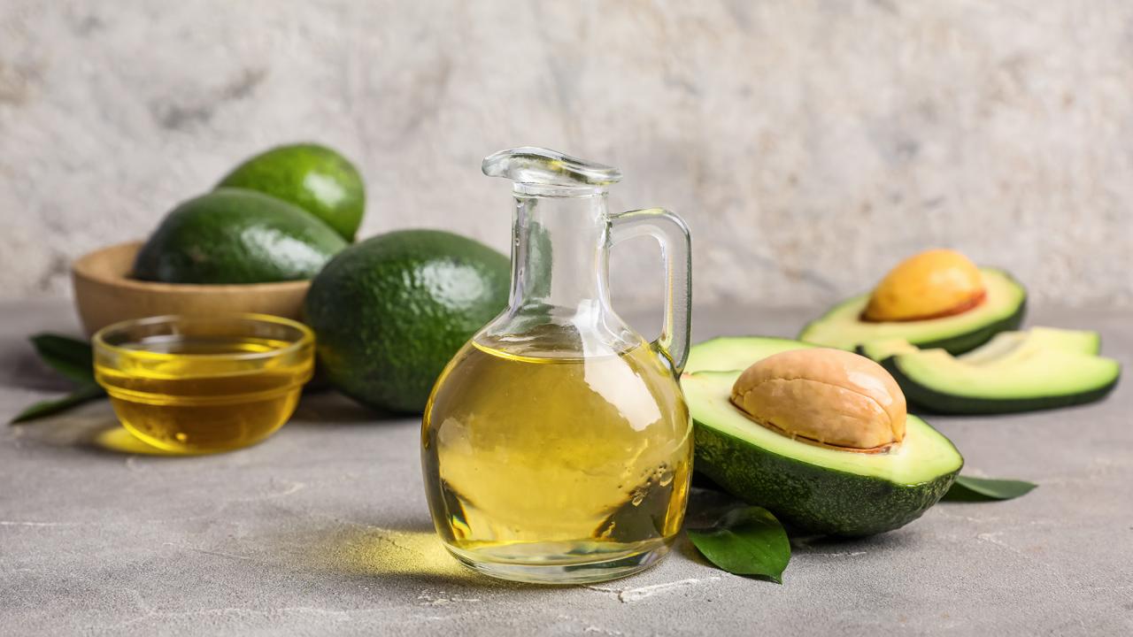 Glass bottle and bowl of golden avocado oil with whole and halved avocados on gray surface