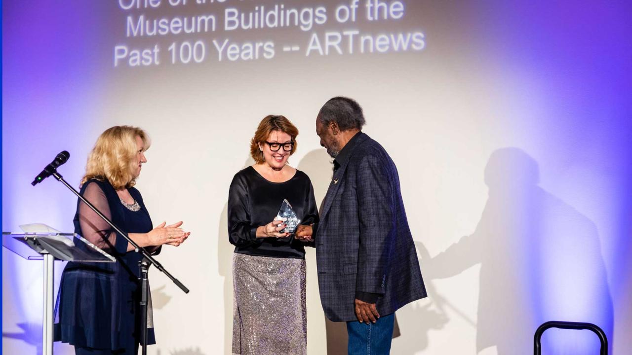 Mike Henderson stands on stage to accept a glass award that reads "Manetti Shrem Museum." Mary Croughan and Rachel Teagle stand opposite Henderson and present the award to him.