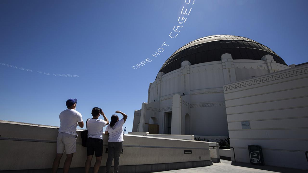 Three people on Griffith Observatory terrace pointing at skywriting over the dome