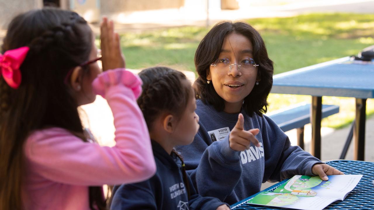 A woman points to the page of a book as two children look on