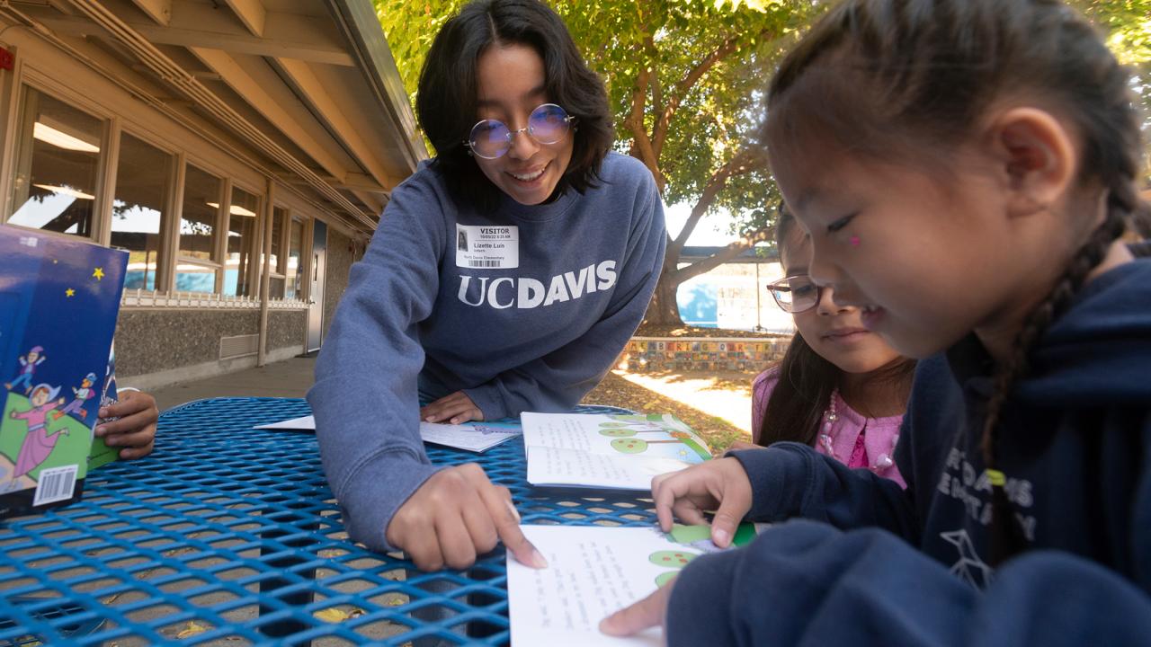 A woman points to the page of a book as two children look on