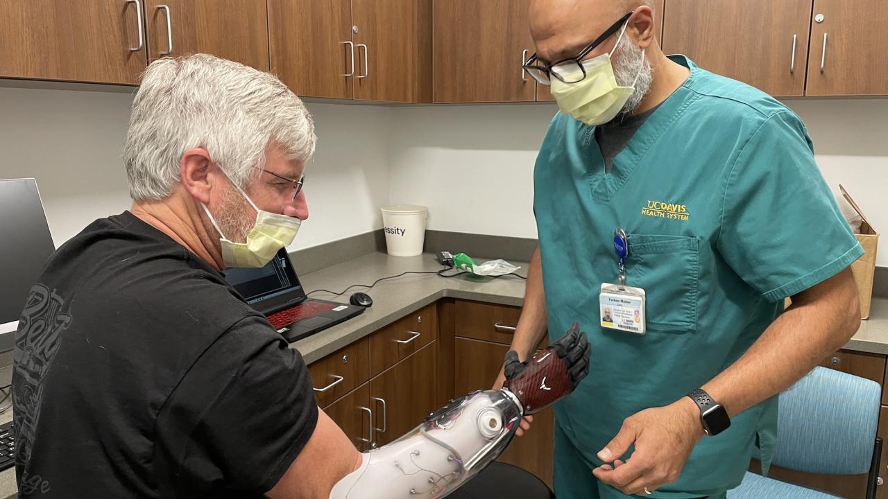 Dave Brockman, a hand amputee, wears a myoelectric prosthesis that looks like a red and black glove. He sits in front of his orthotist who is making sure the fit is correct.