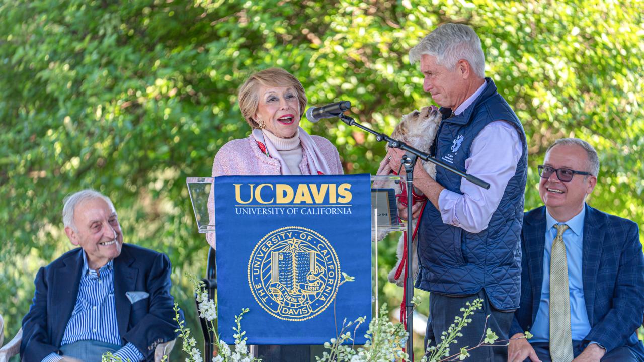 Four people at UC Davis podium outdoors; woman laughing as man speaks, university seal.