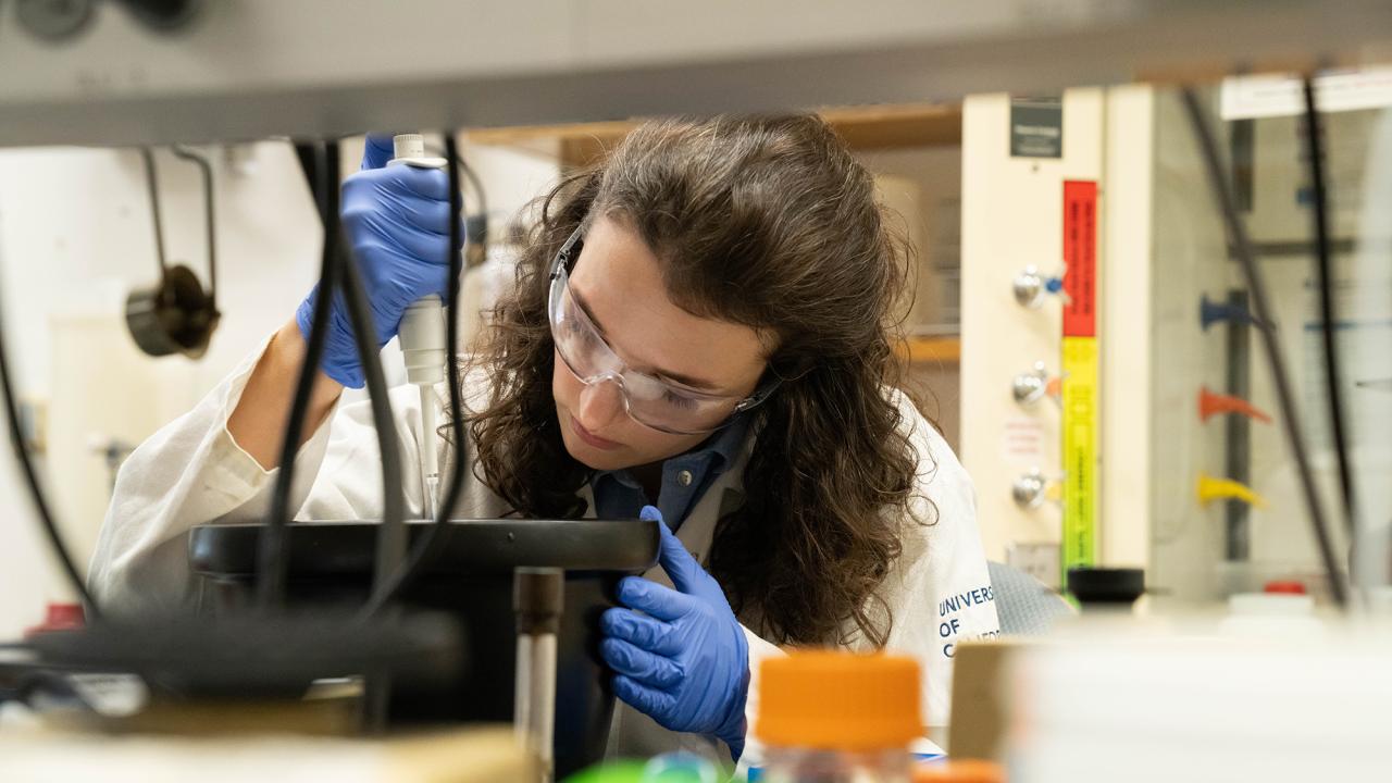 Woman with long brown hair, wearing safety glasses, white lab coat and blue gloves, working at a lab bench. 