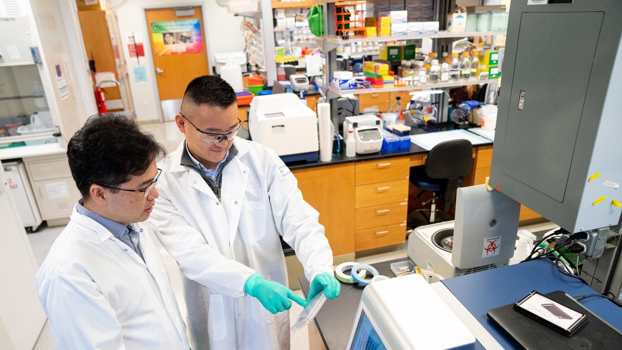 Two men wearing white lab coats in a laboratory setting. One is pointing to a plastic plate. 