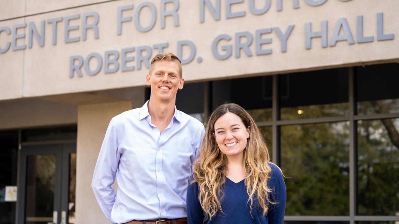 A tall man with blonde hair wearing a light blue shirt stands next to a shorter woman with long brown hair in a dark blue shirt in front of a building entrance. Signage reads Center for Neuroscience Robert D. Grey Hall