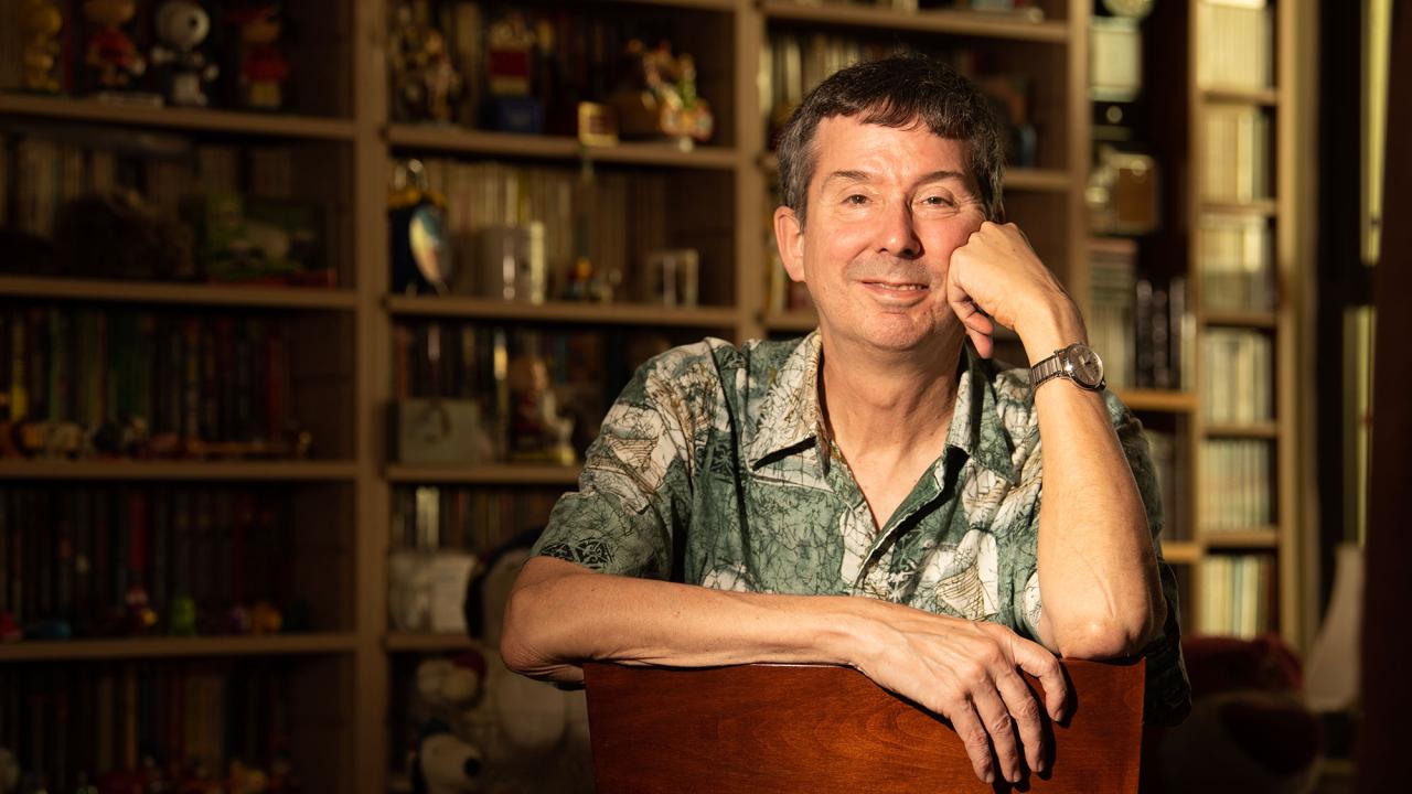 Smiling man in patterned shirt leaning on a chair in warm-lit book-filled room