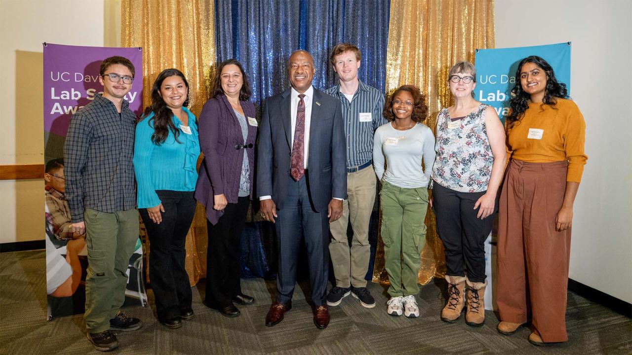 Eight adults standing side-by-side, smiling at indoor event with blue and gold backdrop