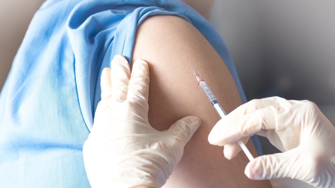 Gloved hands holding a needle and syringe against the bared upper arm of a person wearing a light blue top. 