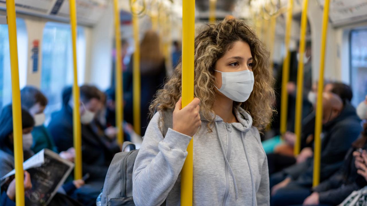 A young woman with long curly brown hair wearing a grey hoodie and face mask while riding light rail. Other passengers in the background also wear masks. 