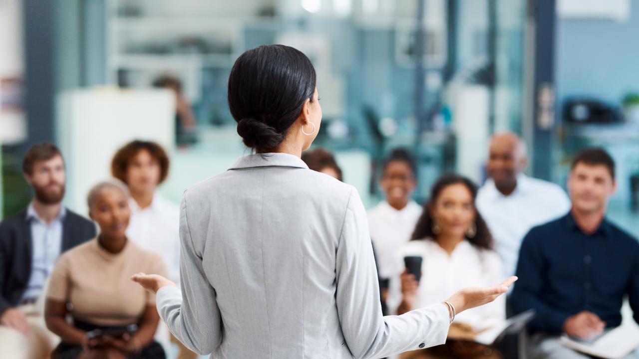 Woman with back to camera stands facing seated group of people listening to her