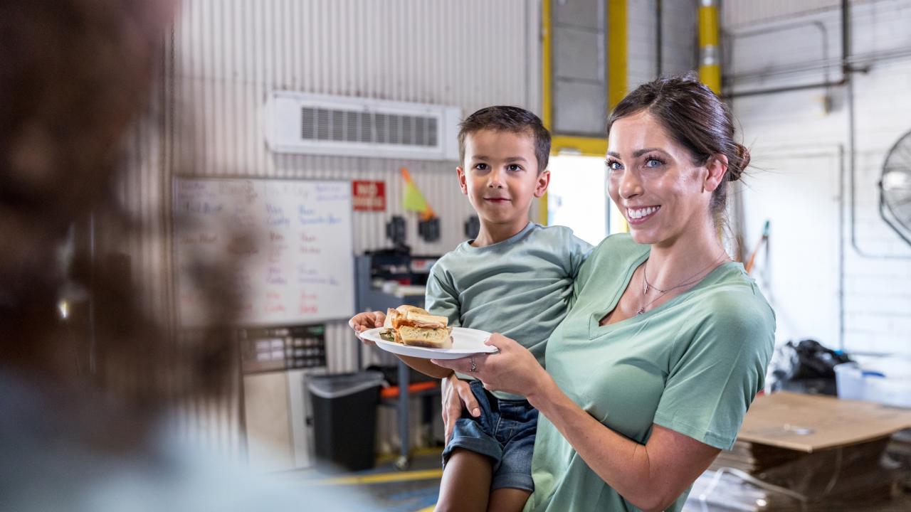 Woman holding child and plate with sandwich at lunch counter.