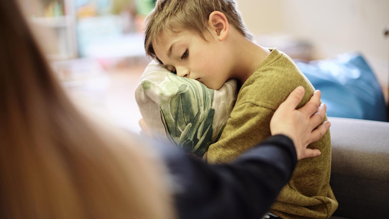 Upset boy hugging pillow on couch while an adult reaches to comfort him.