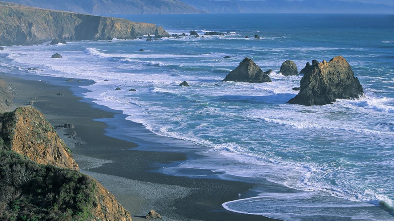 View of rocky coastline and beach to left with island rocks among the surf to right. 