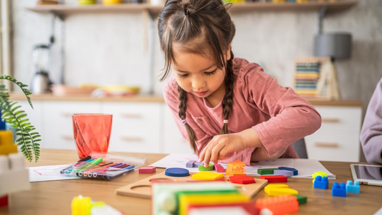 A young girl with braids wearing a pink top concentrates on playing with blocks. 