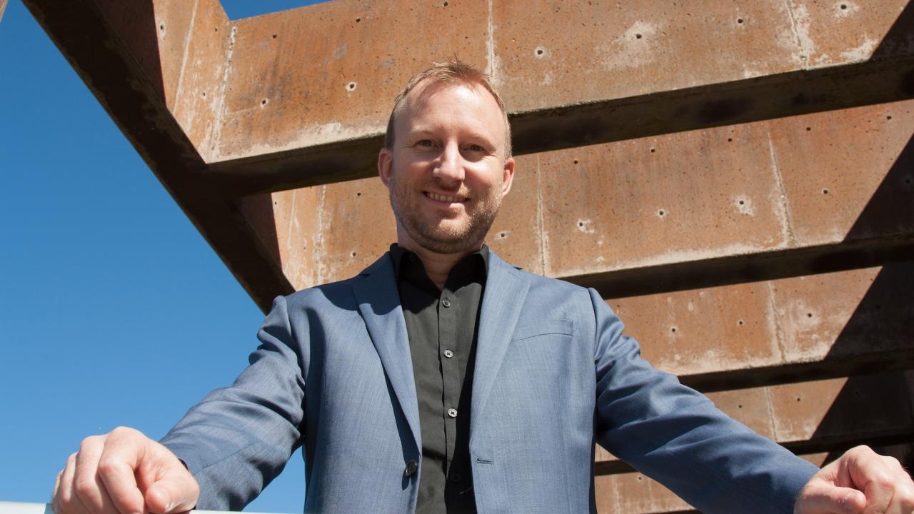 A man in a blue blazer stands confidently on a balcony under a clear blue sky.