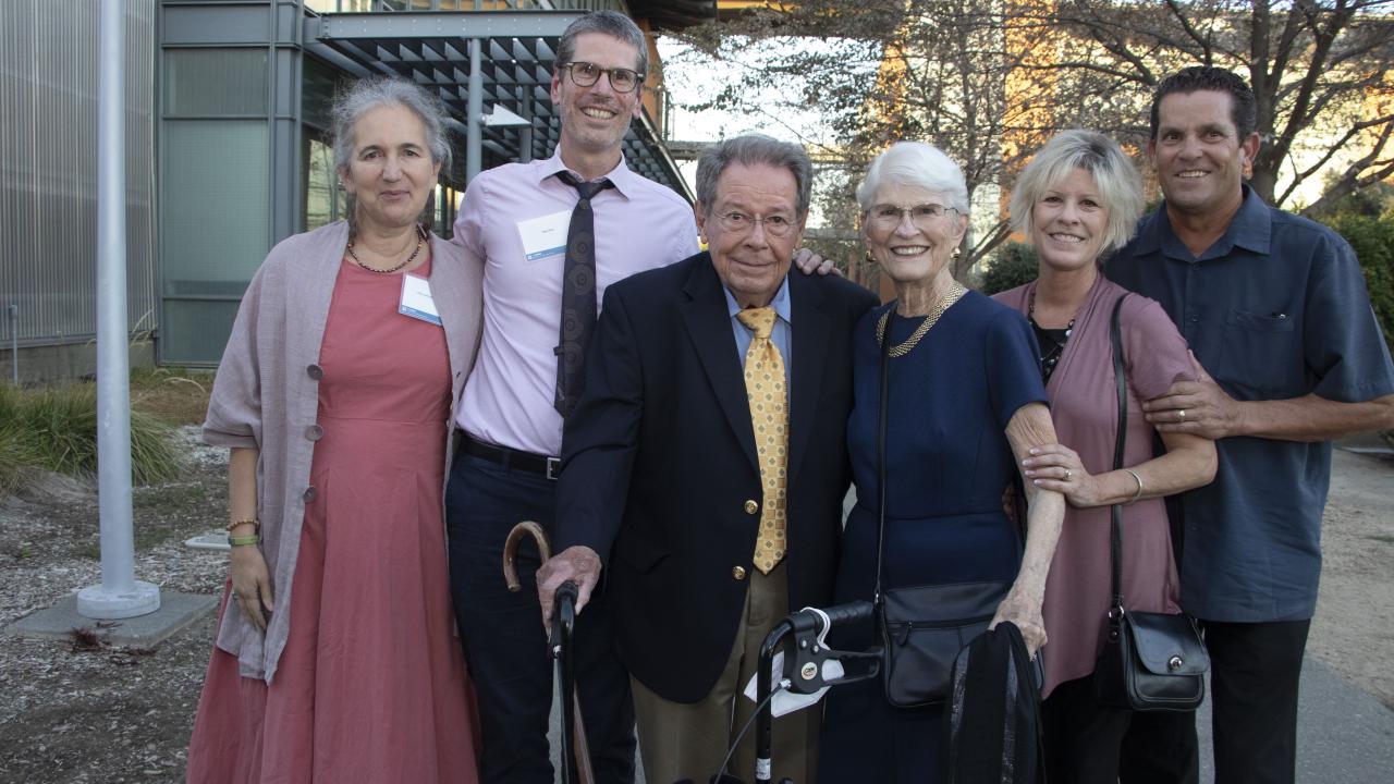 Six smiling adults outdoors, framed by a building a trees, one with a cane, one with a walker