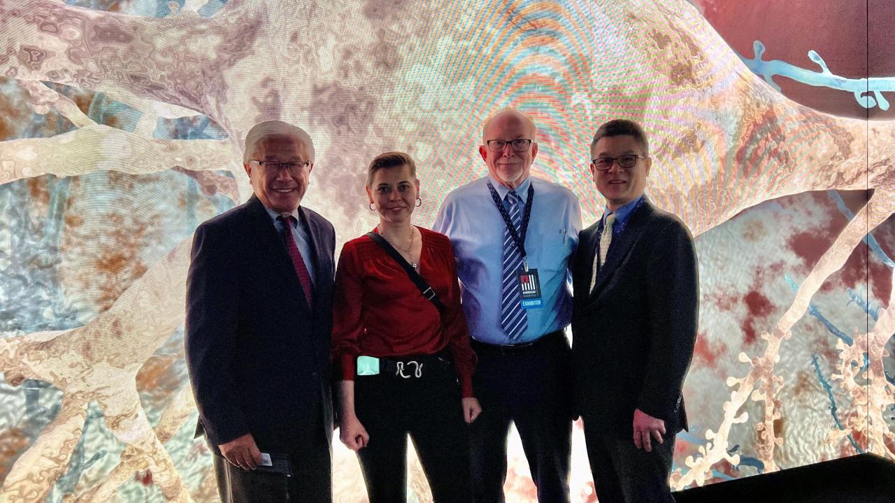 Four people in business attire face the camera. Behind them is a colorful, brightly lit wall display. 