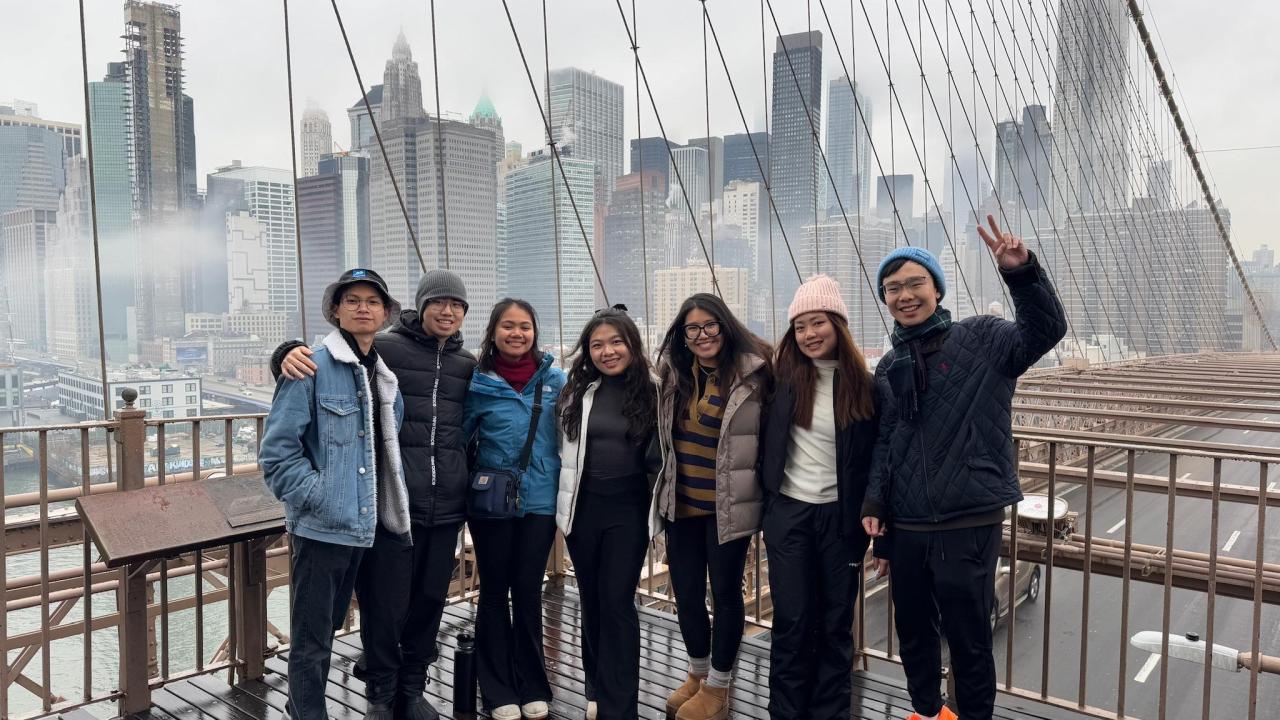 A group of seven young adults stands close together, smiling on the Brooklyn Bridge in New York City. The overcast skyline and tall buildings are visible behind them through the bridge’s cables. Everyone wears winter jackets, hats, and scarves. The bridge's wooden walkway appears wet and one person on the far right flashes a peace sign.