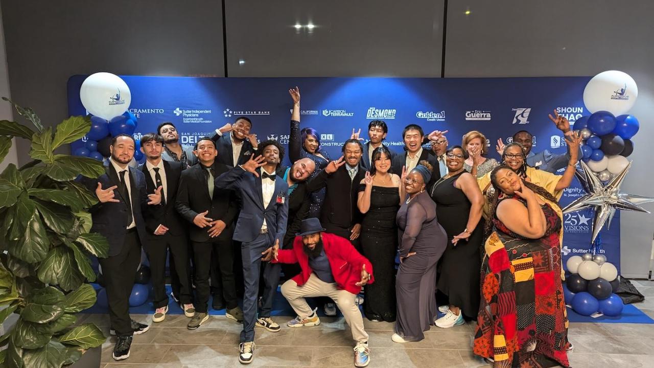 A large group of students and staff pose in formal wear at a blue-carpet event. They stand in front of a branded backdrop with sponsor logos. Many are making fun or celebratory gestures and smiling. Blue and white balloon arrangements frame the scene.