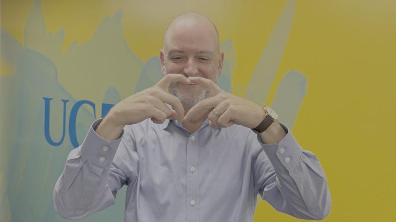 Robert Penman smiles and forms a heart with his hands in front of a colorful blue and yellow UC Davis mural. 