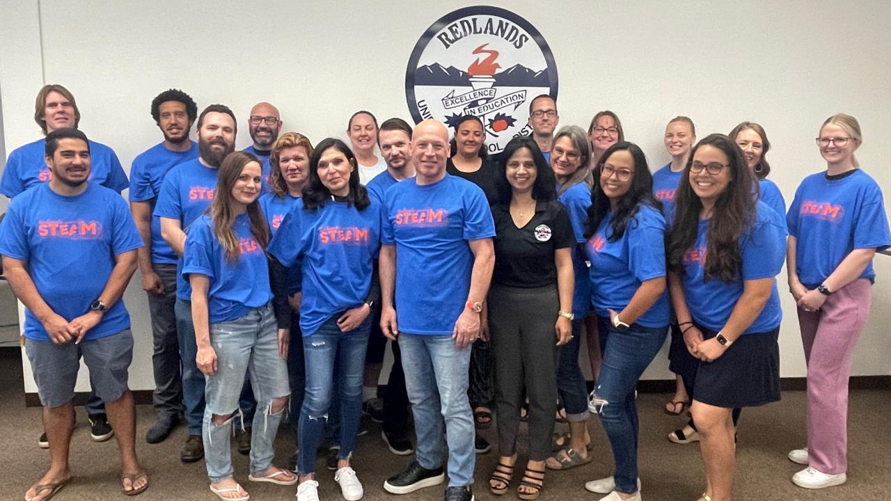 Mixed group of about 20 adults facing camera. Most are casually dressed and wearing blue sweatshirts. A logo on the wall behind them reads "Redlands Excellence in Education"