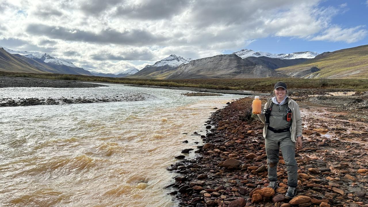 Scientist Taylor Evinger in green jacket and orange pack on rocky riverbank by muddy river and snow-capped mountains
