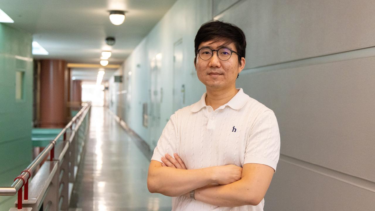 Man in white polo and glasses standing in a long indoor hallway, arms crossed